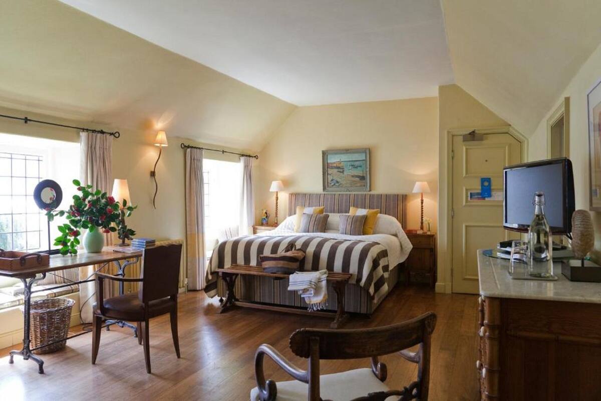 Quarto com cama grande, mesas de cabeceira, escrivaninha com cadeira, janela com cortinas e televisão sobre um móvel.