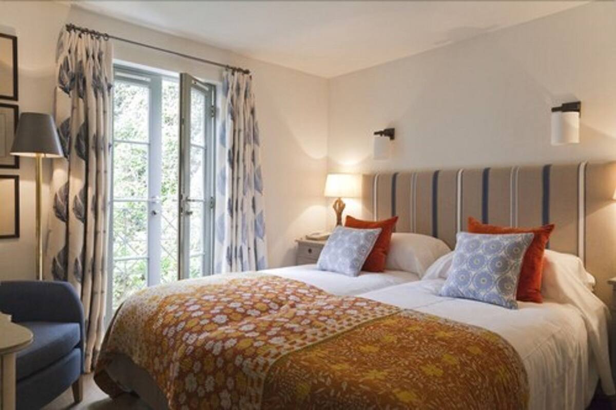 Cama de casal com roupa de cama estampada, duas luminárias de cabeceira e vista para o verde através de uma janela com cortinas.