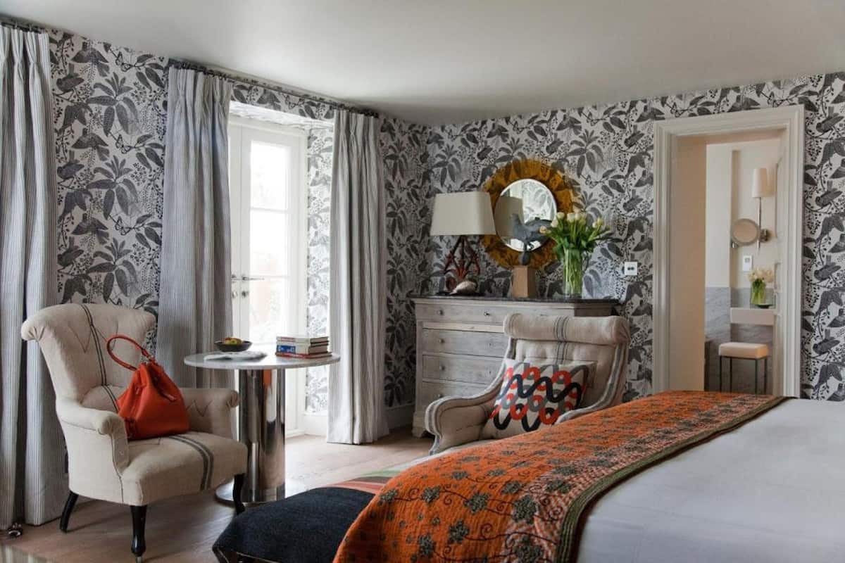 Quarto com cama, cadeira, penteadeira, espelho redondo e mesinha de cabeceira com abajur.