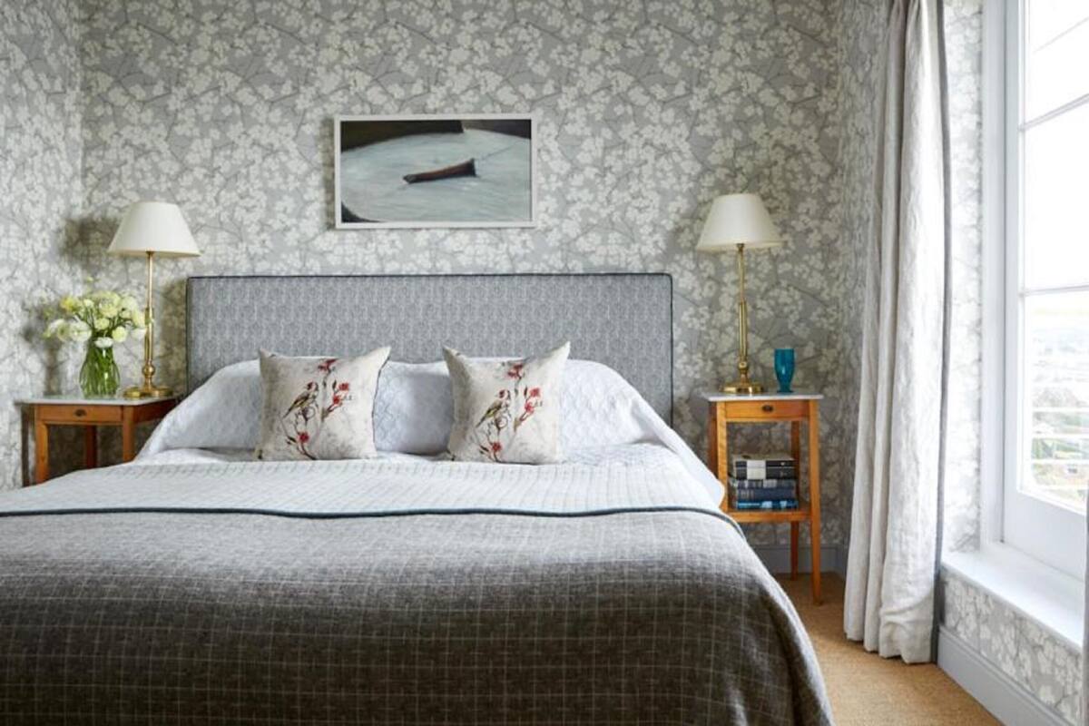 Quarto bem arrumado com uma cama grande, duas mesas de cabeceira com abajures, um quadro na parede e uma janela com cortinas.