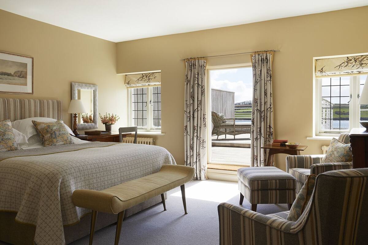 Quarto de hotel com uma cama grande, um banco, duas poltronas, uma mesa, uma cadeira, janela com cortinas e vista para o exterior.