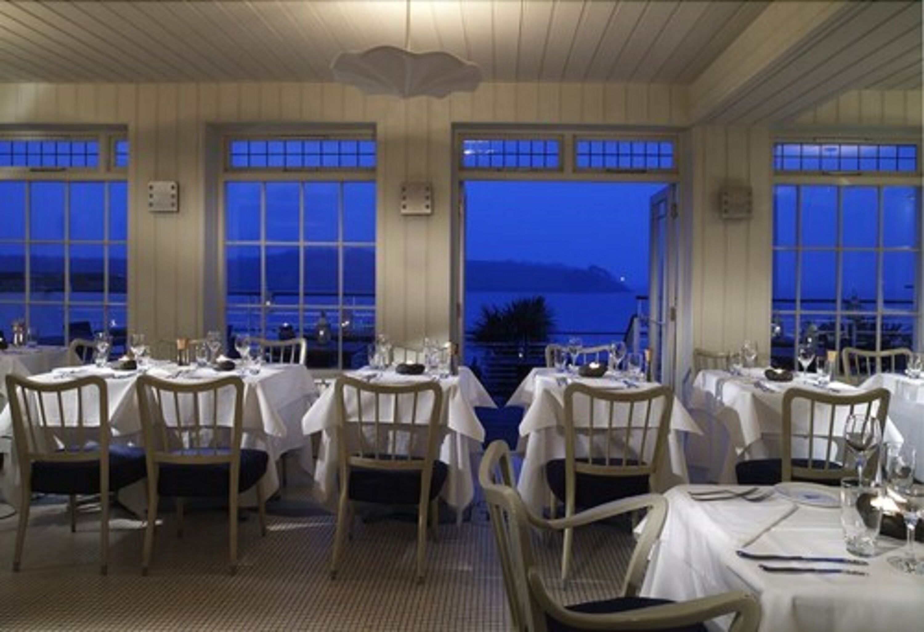 Un restaurante con mesas dispuestas para comer, ventanas amplias y vistas al mar.