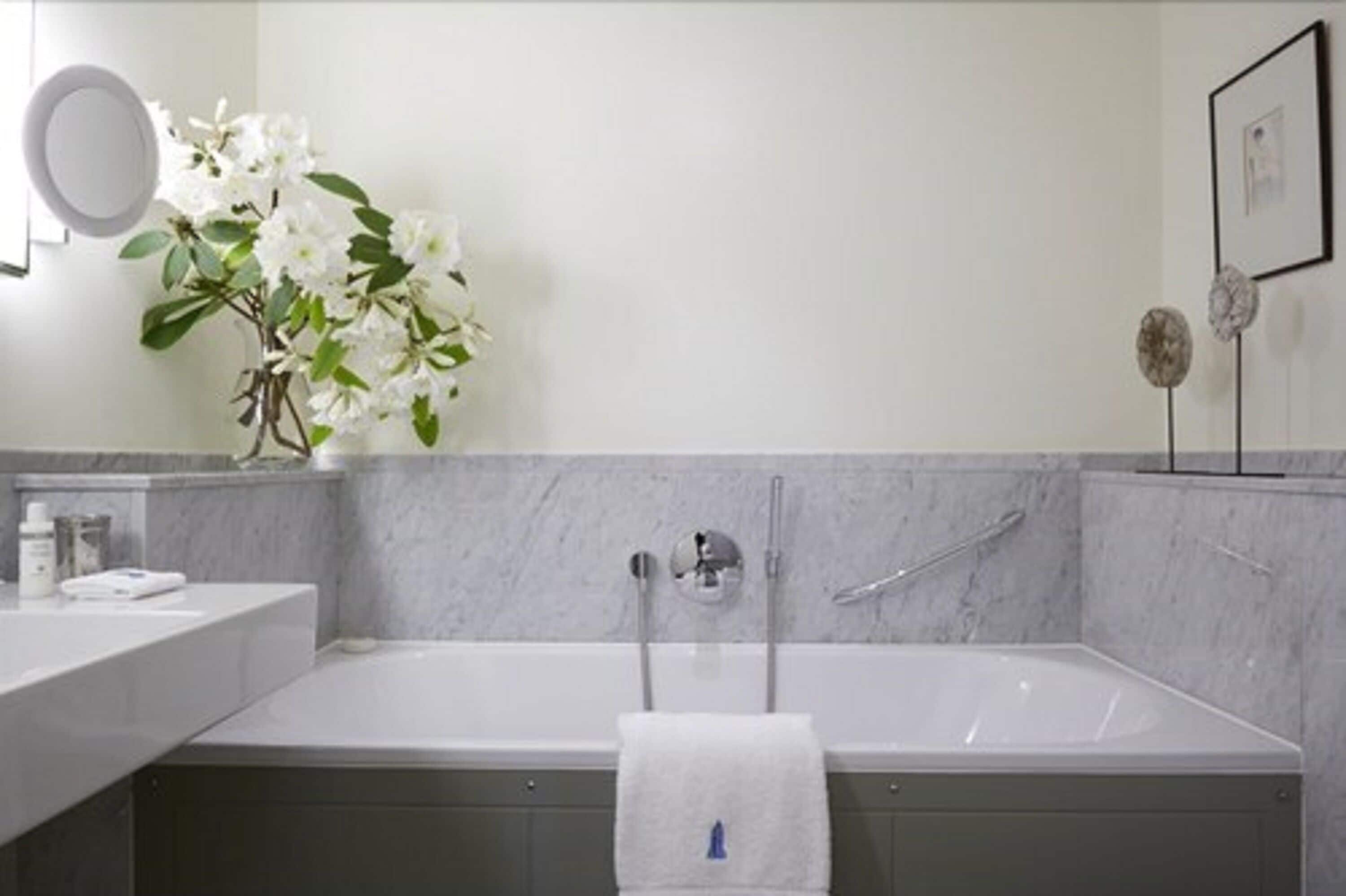 Un baño moderno con pared de mármol, una bañera blanca, un espejo redondo y un florero con flores blancas.