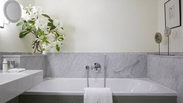 Un baño moderno con pared de mármol, una bañera blanca, un espejo redondo y un florero con flores blancas.