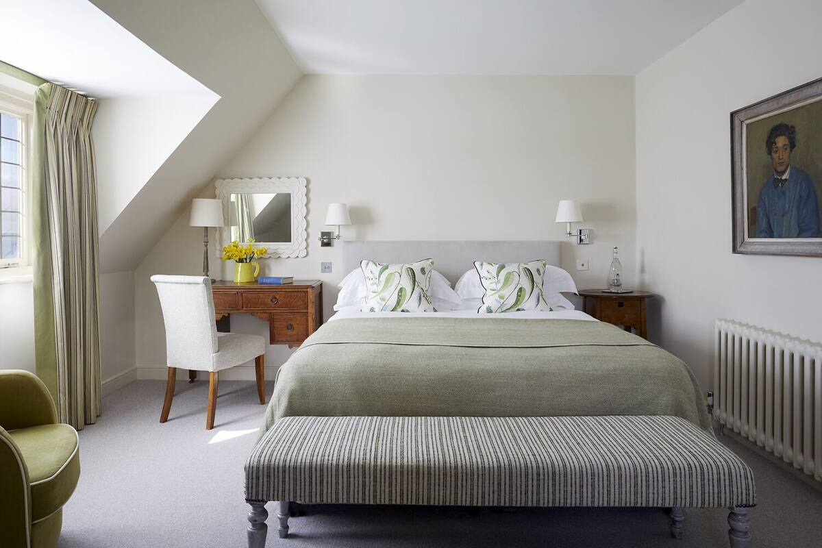 Quarto de hotel com uma cama grande, uma escrivaninha, uma cadeira, cômoda, espelho, quadro e radiador.