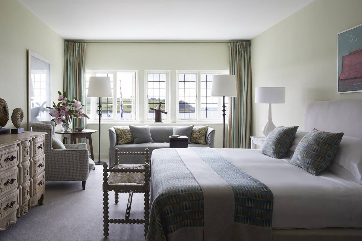 Quarto de hotel com uma cama grande, um sofá, uma cadeira, cômoda e vista para o mar através de grandes janelas.