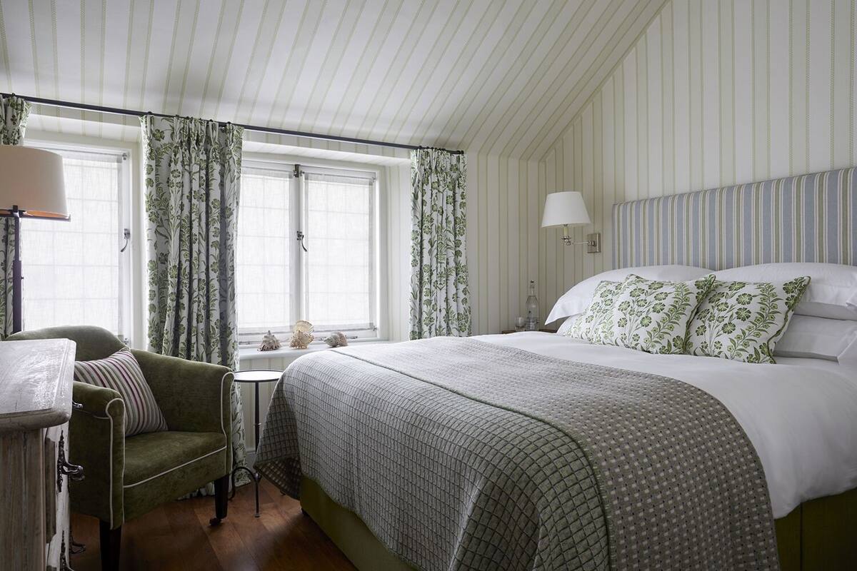 Quarto de hotel com cama grande, uma cadeira, uma mesa pequena e cortinas.