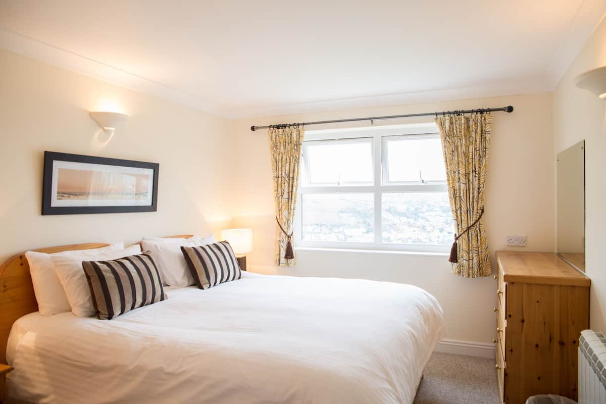 Een slaapkamer met een groot bed, een nachtkastje, een lamp, een raam met gordijnen, een lijst met een afbeelding aan de muur en een houten dressoir.