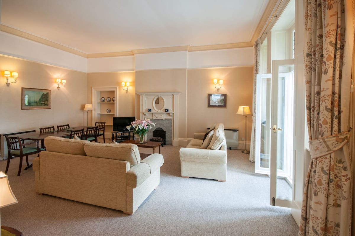 Een woonkamer met een bank, fauteuils, een open haard en een eethoek met stoelen en een tafel.