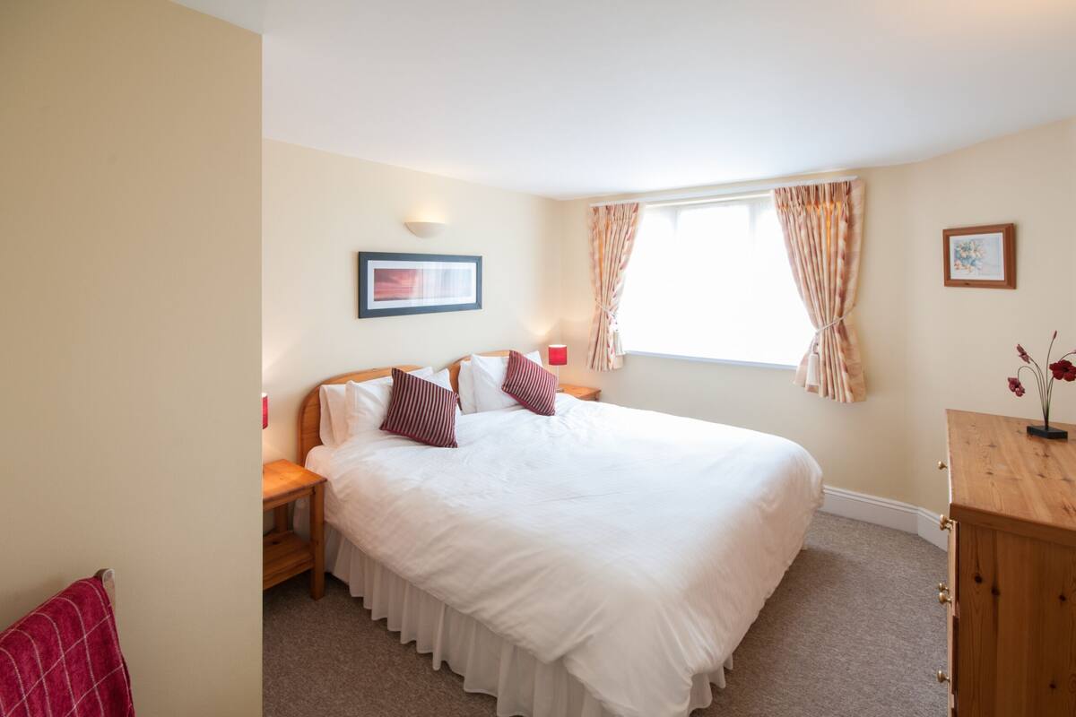 Een netjes ingerichte slaapkamer met een groot bed, een nachtkastje, een houten dressoir en een raam met gordijnen.