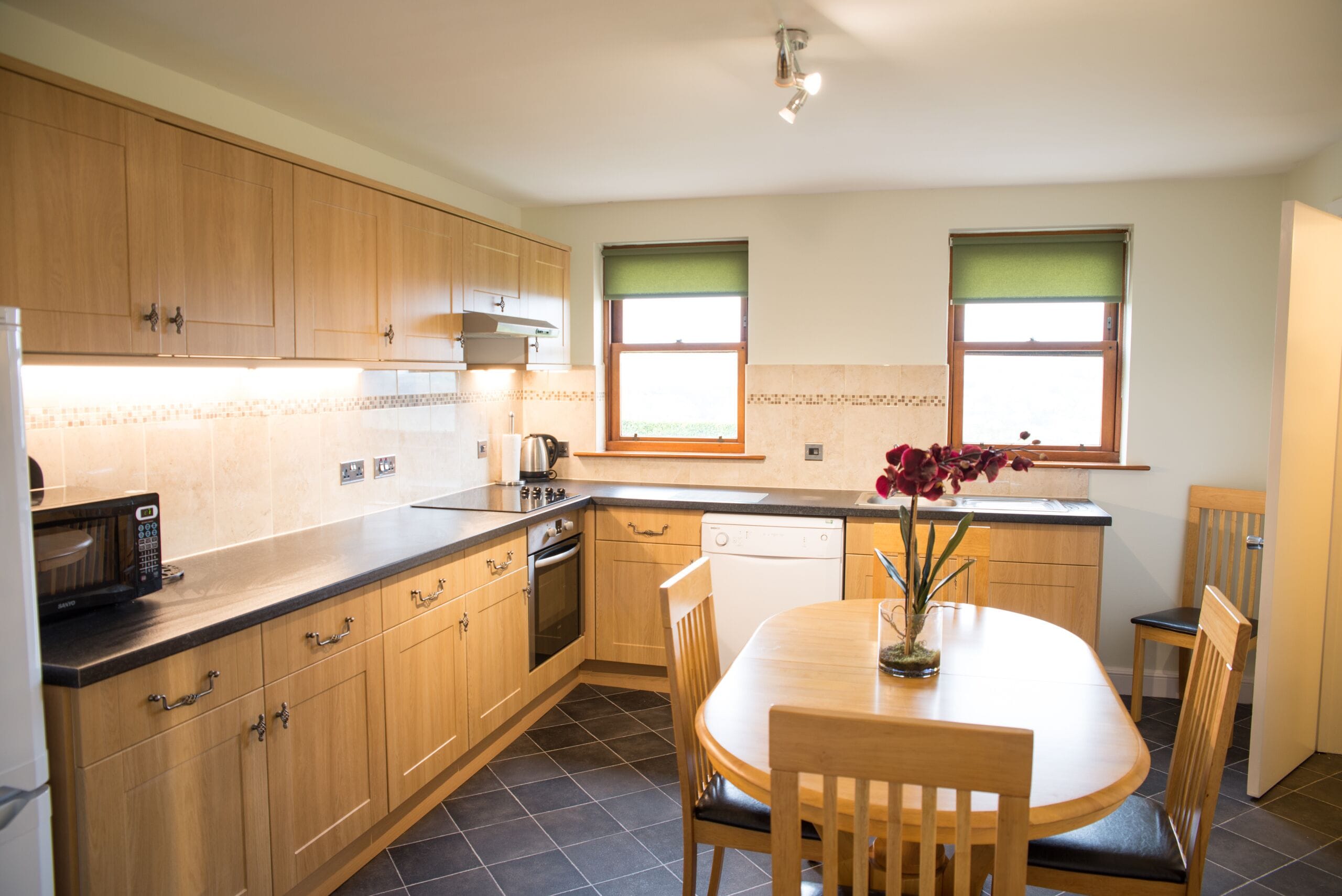 A modern kitchen with wooden cabinets, a microwave, an oven, and a dishwasher. There is a dining table with chairs and a vase of flowers.