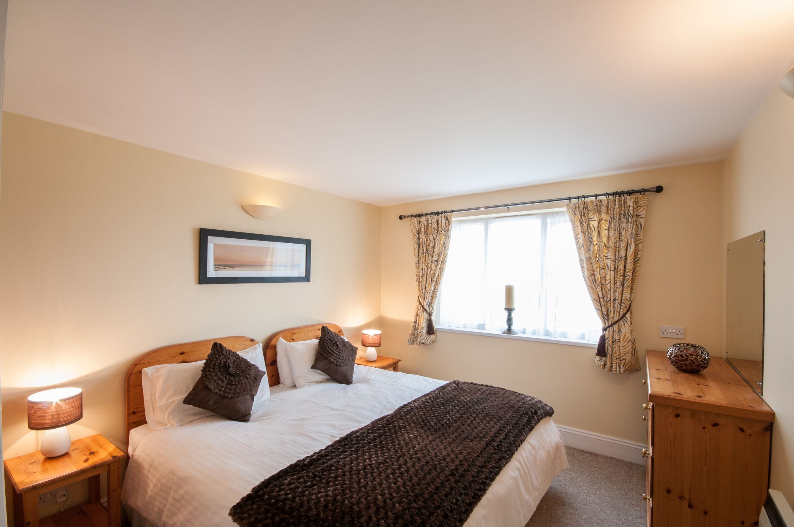 A bedroom with a bed, bedside lamps, a window with curtains, a framed picture on the wall, and a wooden dresser.