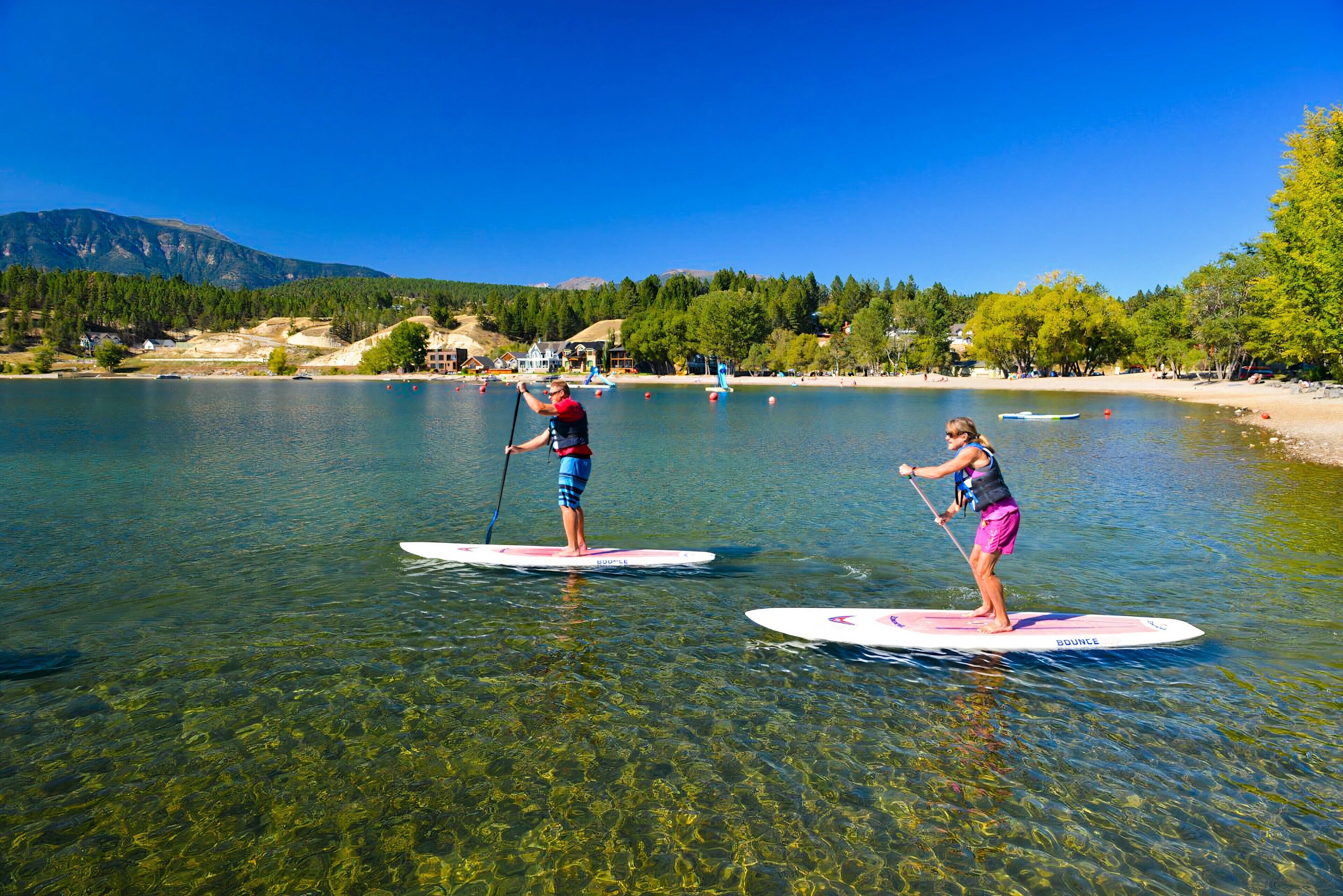 Two people paddleboarding on a clear lake with a mountainous backdrop.