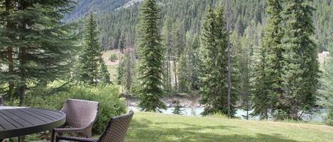 An outdoor seating area with a table and chairs, overlooking a river and forested mountains.