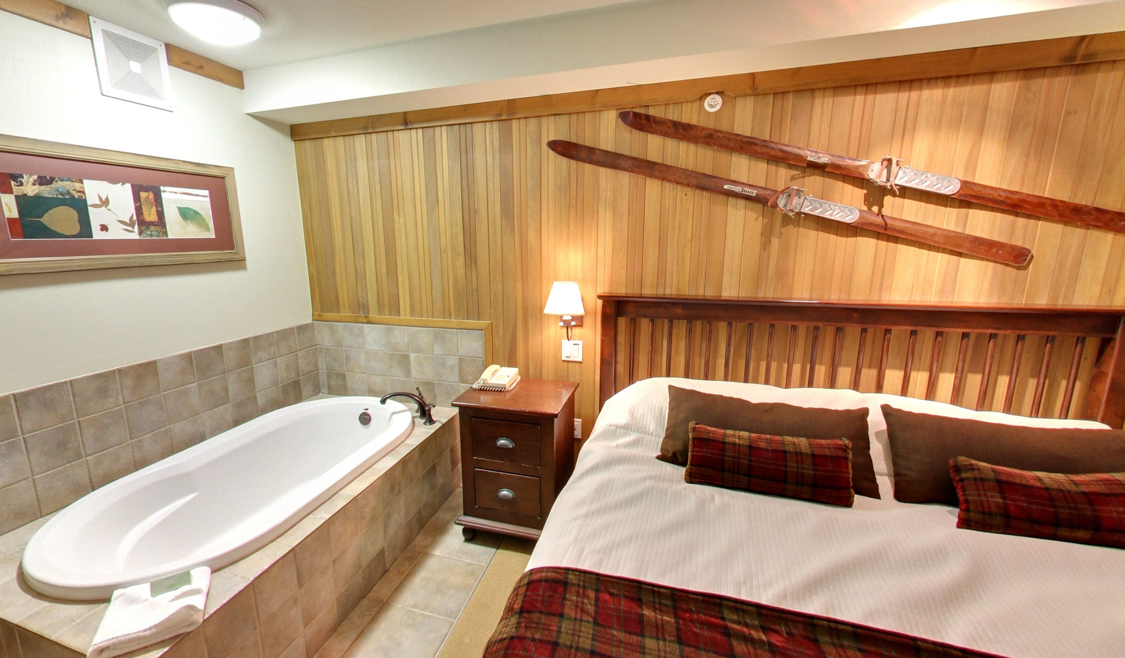 Une chambre d’hôtel avec une tête de lit en bois, un grand lit avec une couette à carreaux, une table de chevet avec une lampe et un porte-skis fixé au mur.