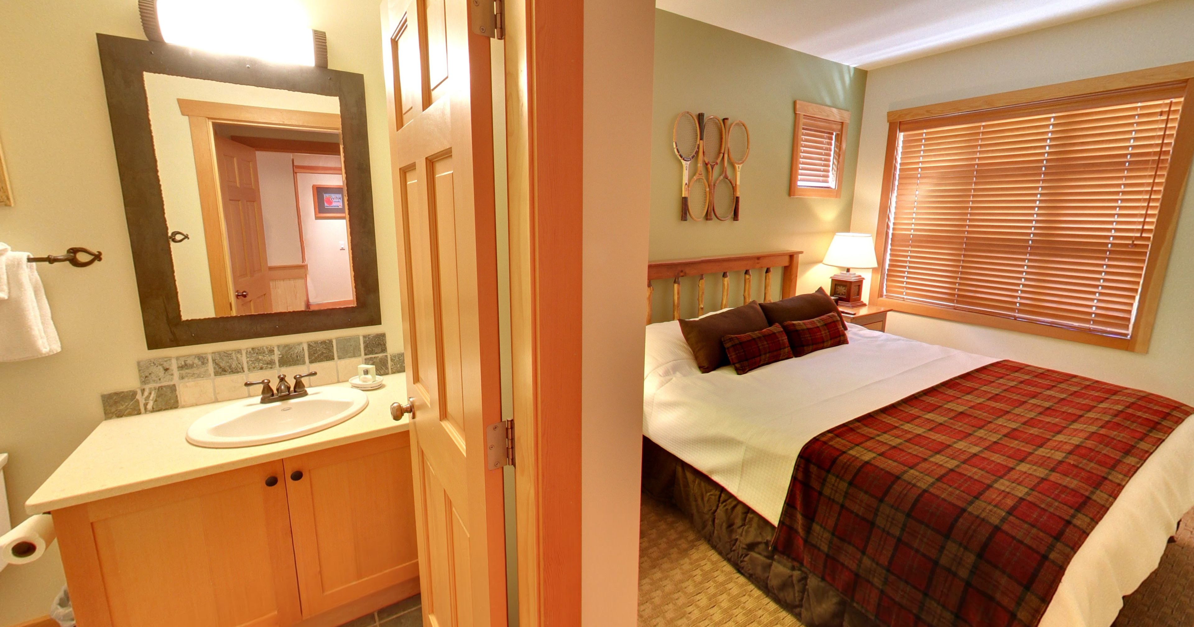 A bathroom with a sink, mirror, and toilet. The room has a wooden door and a window with blinds.