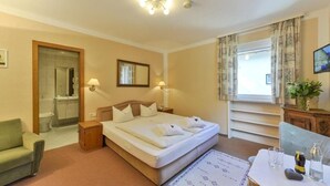 A hotel room with a bed, bedside tables, a sofa, a TV, a window with curtains, and a bathroom visible through an open door. - Gasthof Alpenhof (Mariastein)