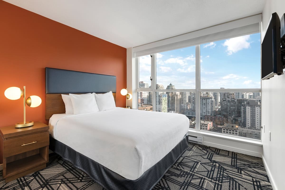 A hotel room with a large bed, a nightstand with a lamp, and a view of a cityscape through a large window.