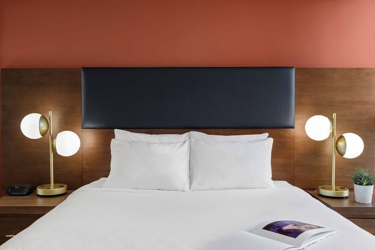 A neatly made bed with white linens, two bedside lamps with round shades, a dark headboard, and a small potted plant on the nightstand.