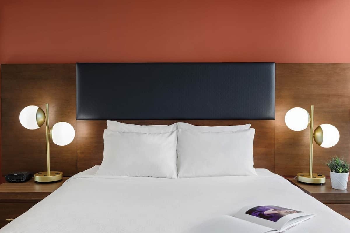 A neatly made bed with white linens, two bedside lamps with round shades, a dark headboard, and a small potted plant on the nightstand.