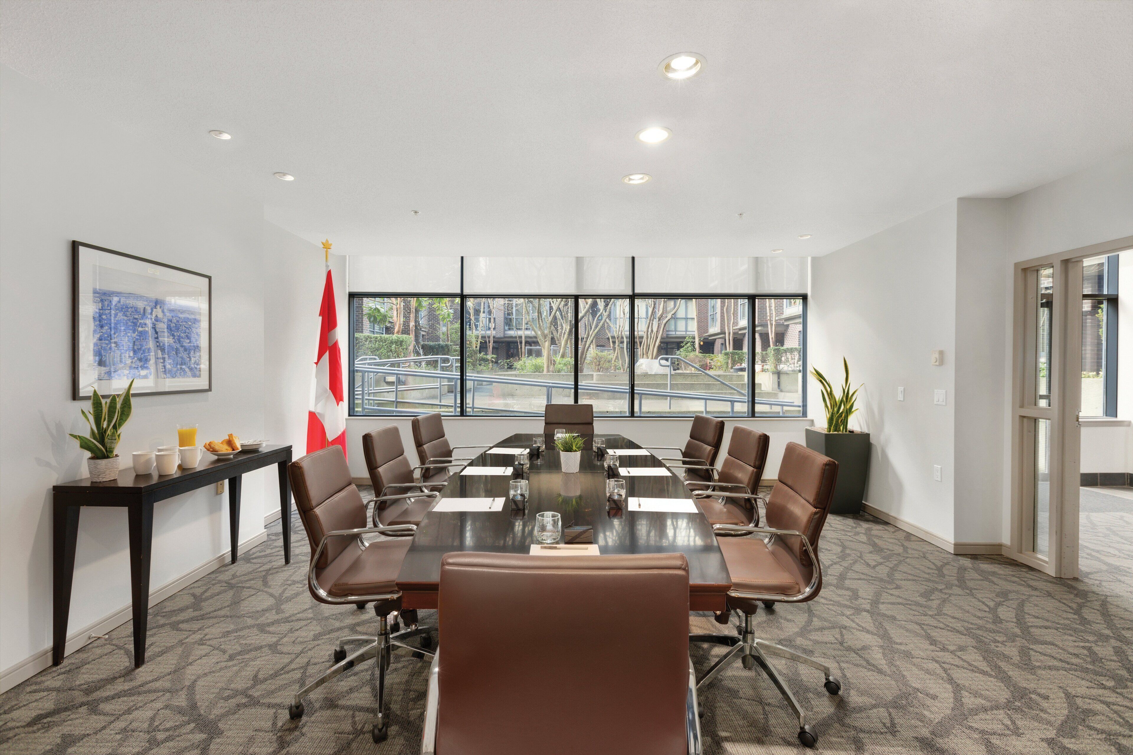 A conference room with a long table, brown chairs, a flag, a framed picture, and a window view.