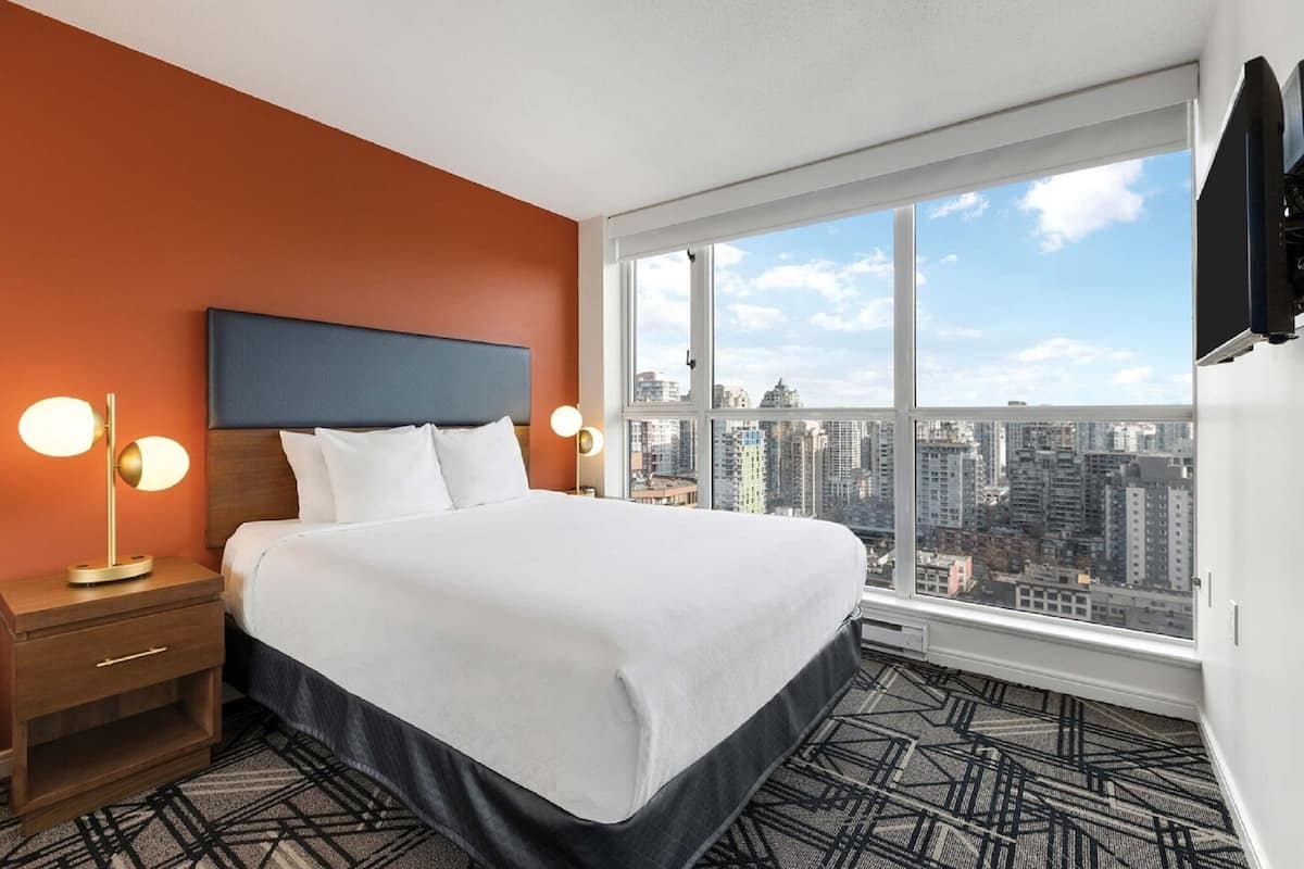A hotel room with a bed, nightstand, lamp, and a large window with a city view.