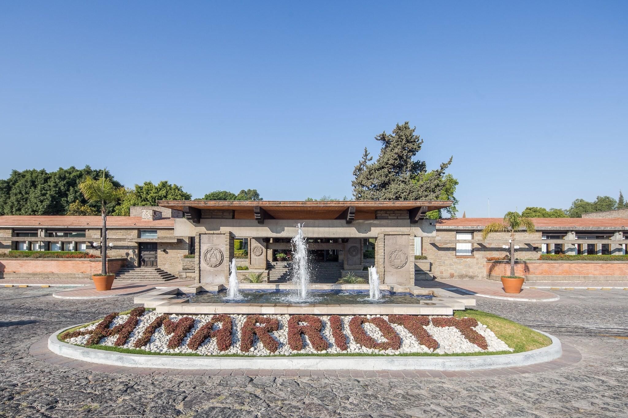 Ein Hotel Eingang mit einem Brunnen, einem Schild mit der Aufschrift 'MARRIOTT' und einem begrünten Bereich mit Bäumen und Sträuchern.