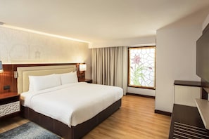 Quarto de hotel com uma cama grande, televisão, janela com cortinas e parede com padrão decorativo.