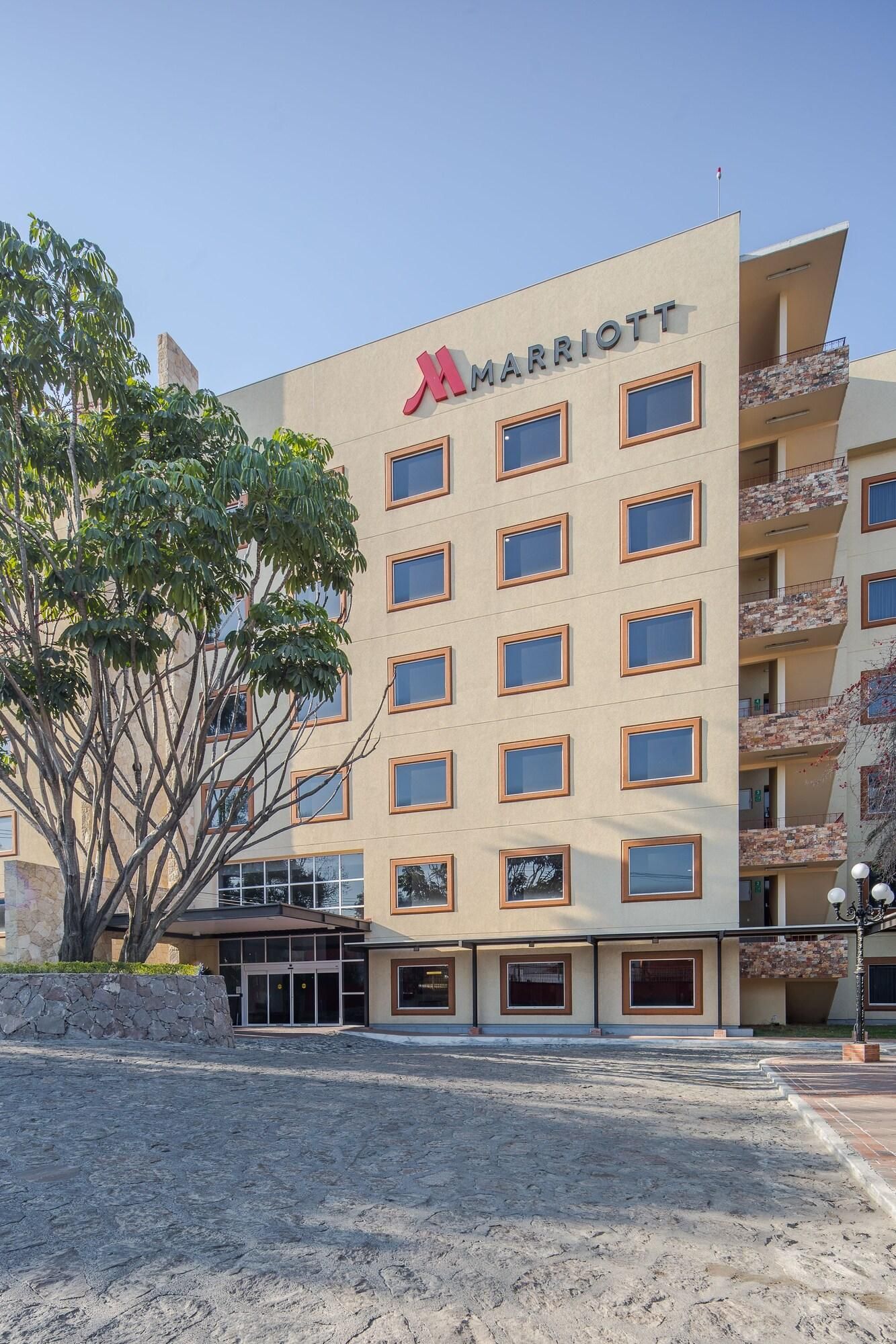 Un hôtel à plusieurs étages, reconnaissable à son logo Marriott bien visible, à sa façade en pierre et à son entrée agrémentée d’un aménagement paysager.