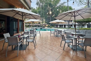 Ein Außenbereich mit Terrasse, Tischen, Stühlen, Sonnenschirmen und einem Swimmingpool.