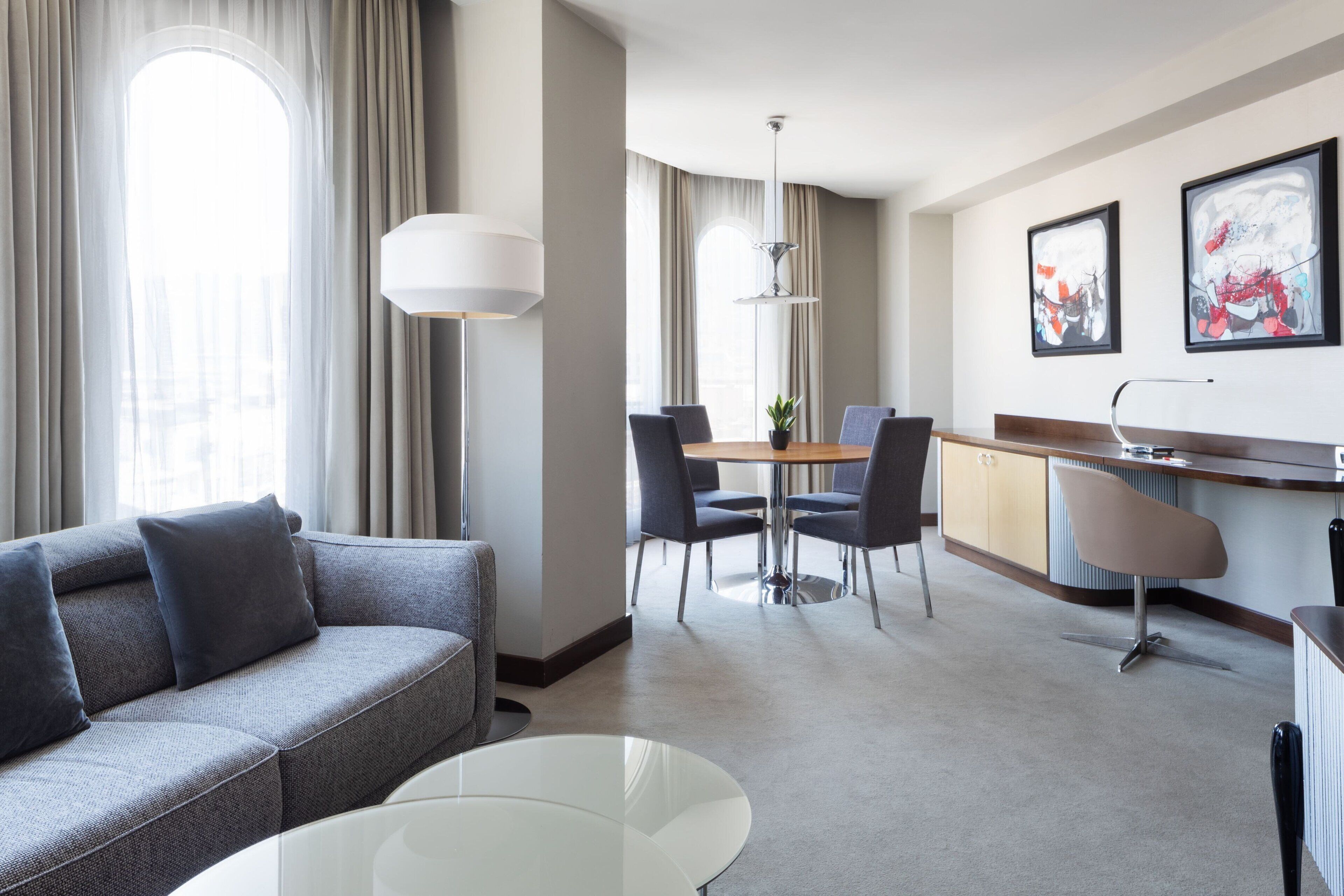 A modern hotel room with a grey sofa, a glass coffee table, a dining area with a table and chairs, a kitchen area with a counter and cabinets, and two framed artworks on the wall.