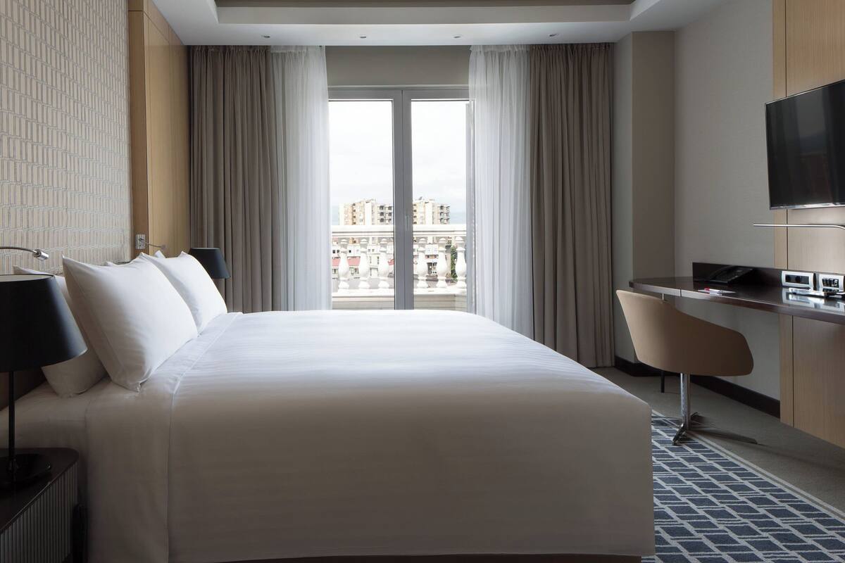 Une chambre d’hôtel avec un grand lit, un bureau, une chaise, une télévision et une vue sur les bâtiments par la fenêtre.