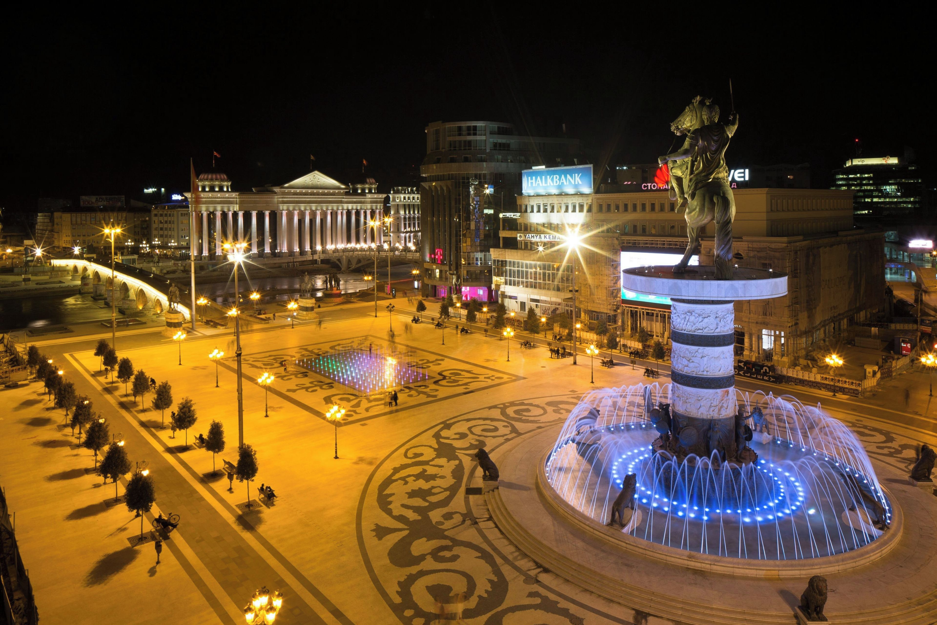 Una plaza urbana nocturna con una fuente central, edificios circundantes y un puente.