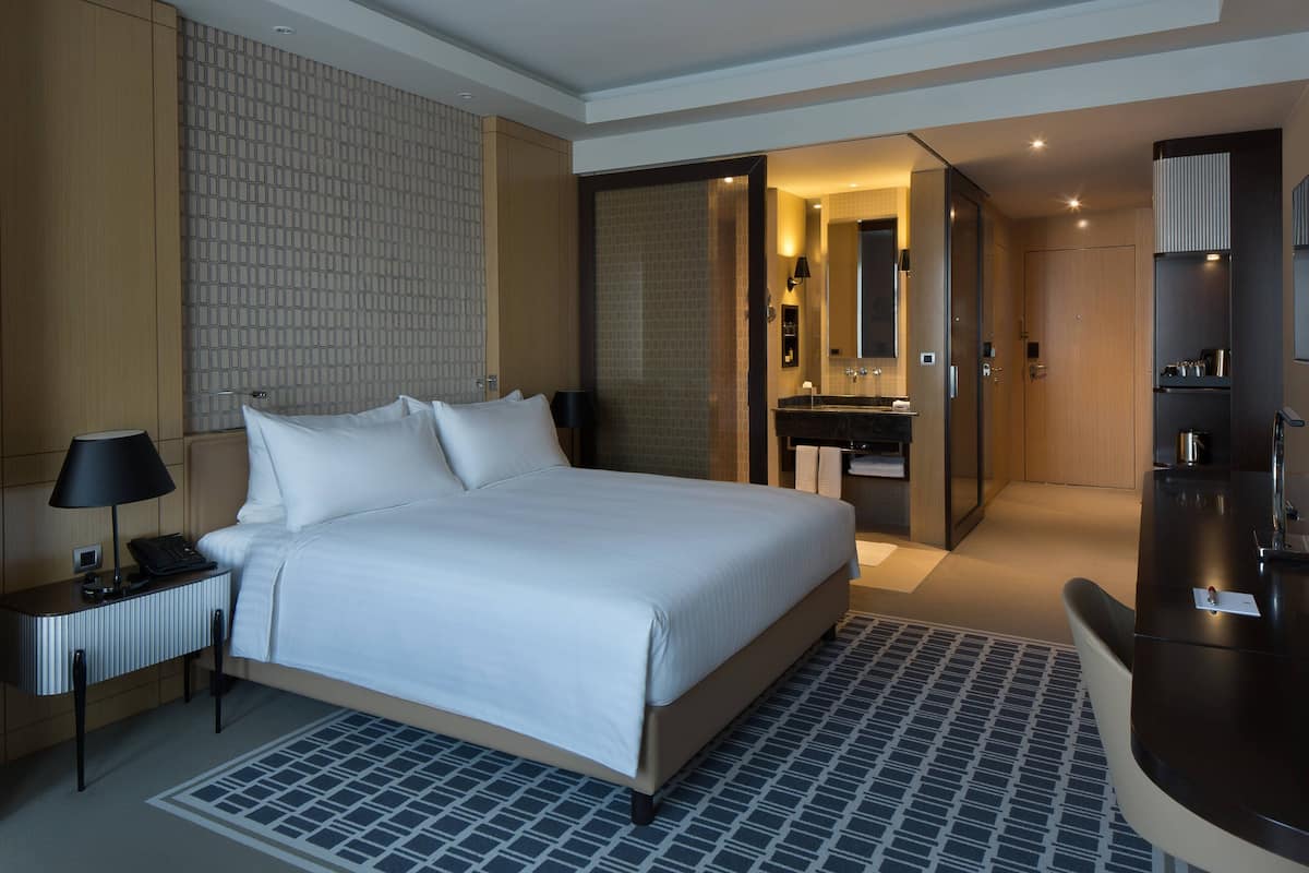 Une chambre d’hôtel avec un grand lit, une table de chevet avec une lampe, une salle de bain avec un miroir et un lavabo, et un bureau avec une chaise.