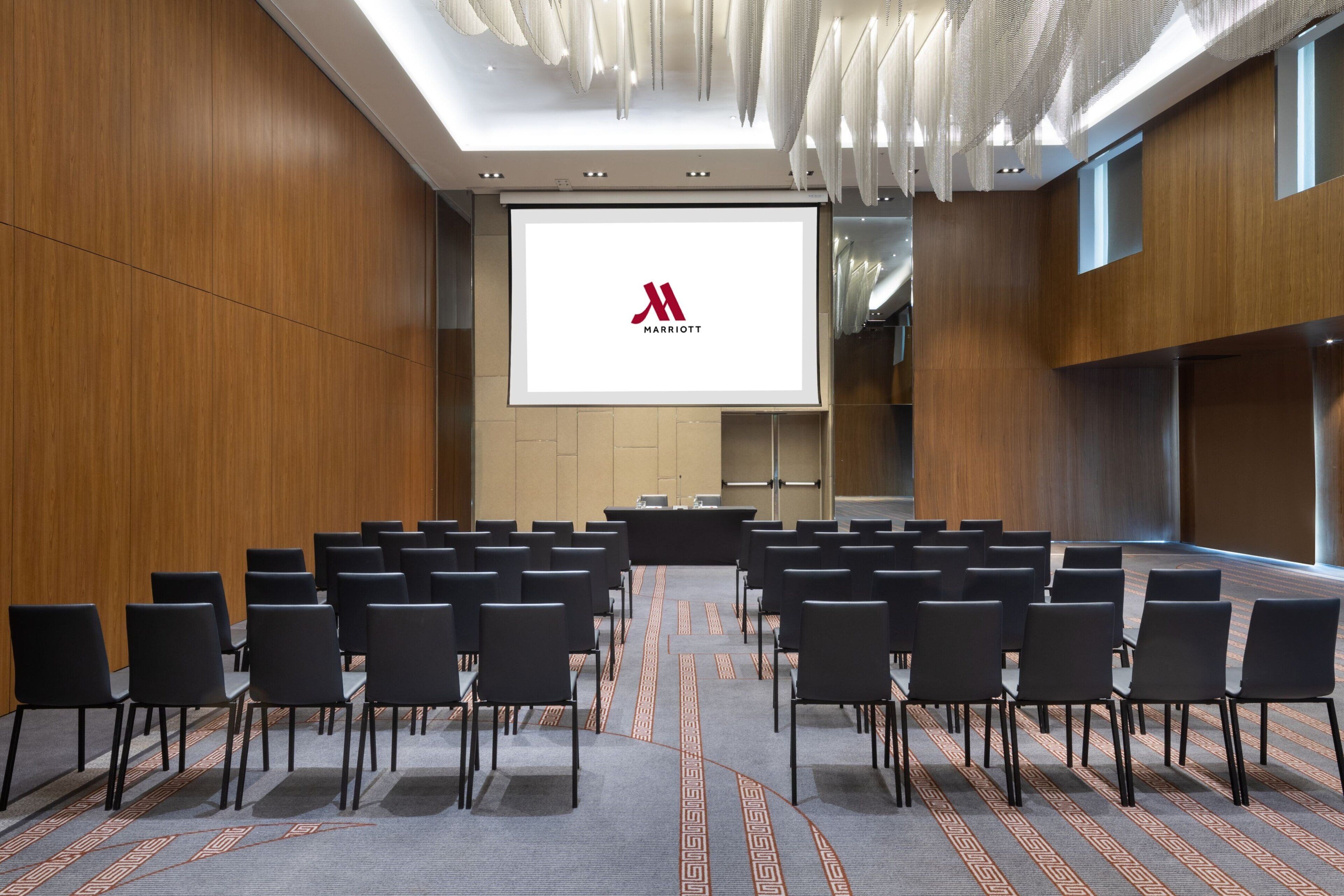 A conference room with rows of black chairs, a large screen displaying the Marriott logo, and a modern ceiling design.