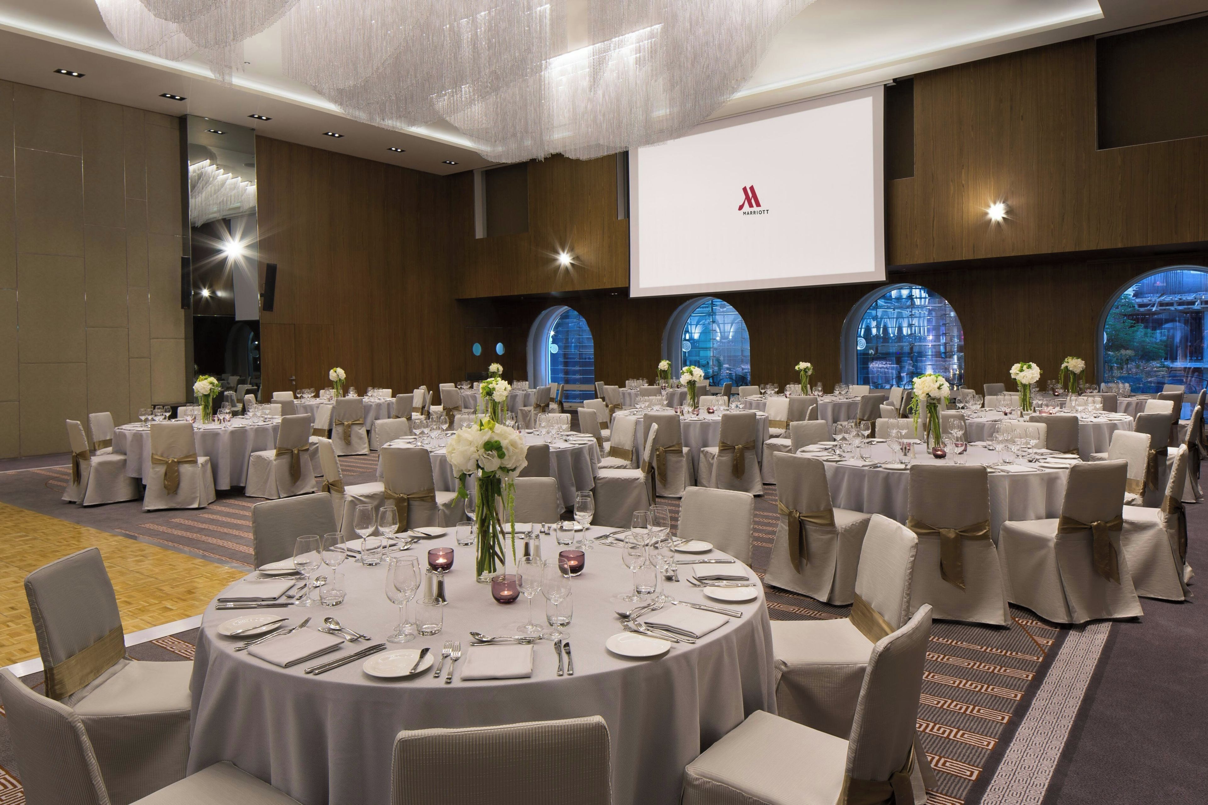 Une grande salle de conférence dotée de plusieurs tables rondes, chacune recouverte d’une nappe blanche et agrémentée de chaises et de compositions florales.