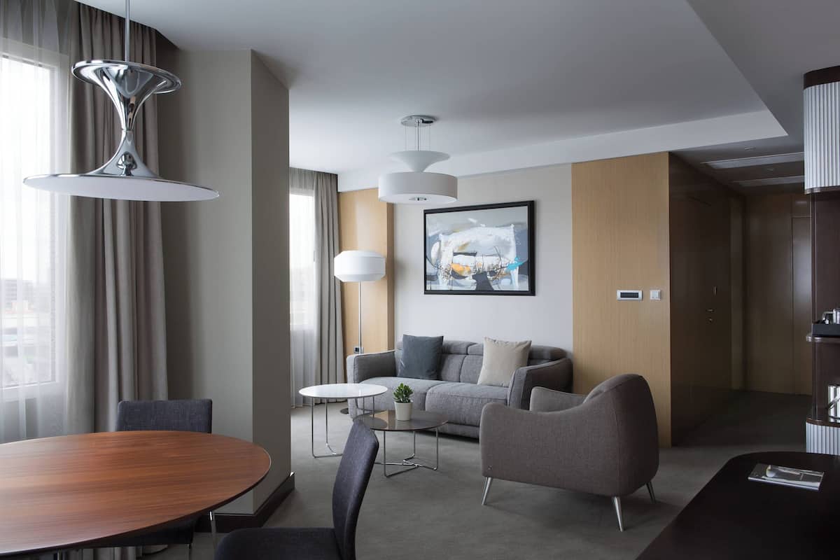 Une chambre d’hôtel moderne avec une table à manger en bois, un canapé gris, une petite table ronde et une grande fenêtre avec des rideaux.