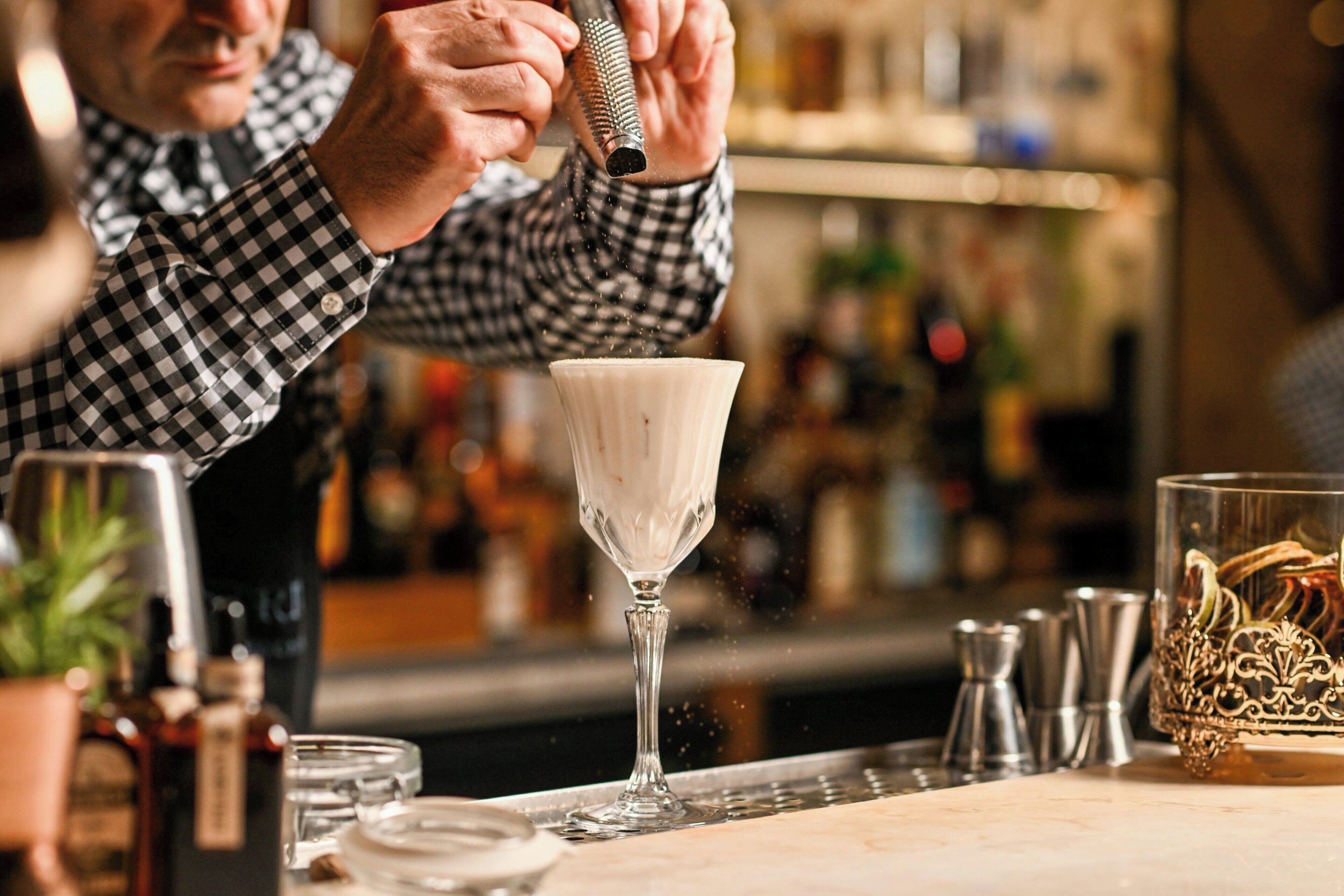A bar setting with a bartender preparing a drink, a glass on the counter, and various bar tools and bottles in the background.