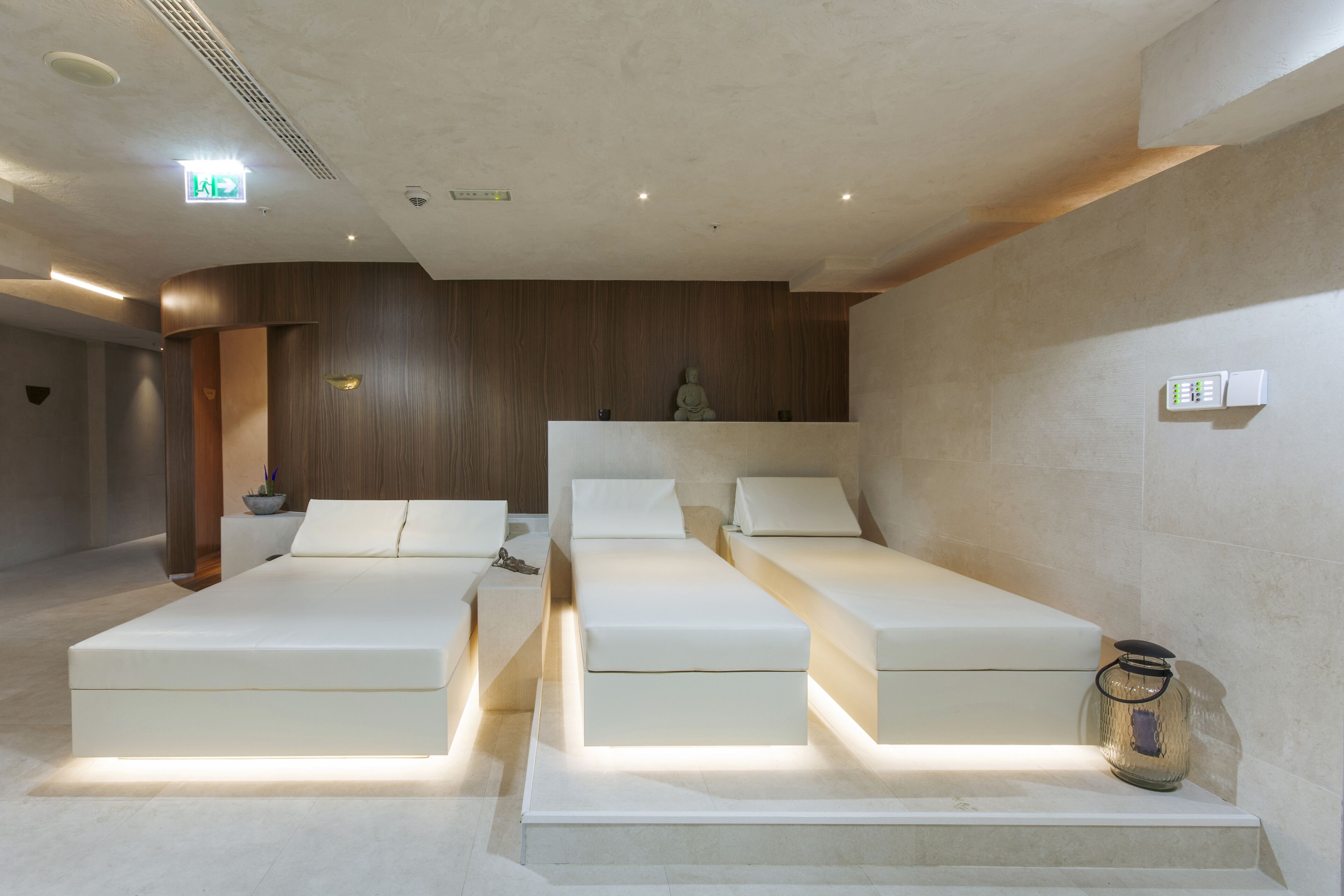 Une salle de spa moderne avec trois lits blancs disposés en rangée, un mur en bois et une petite statue posée sur une étagère.