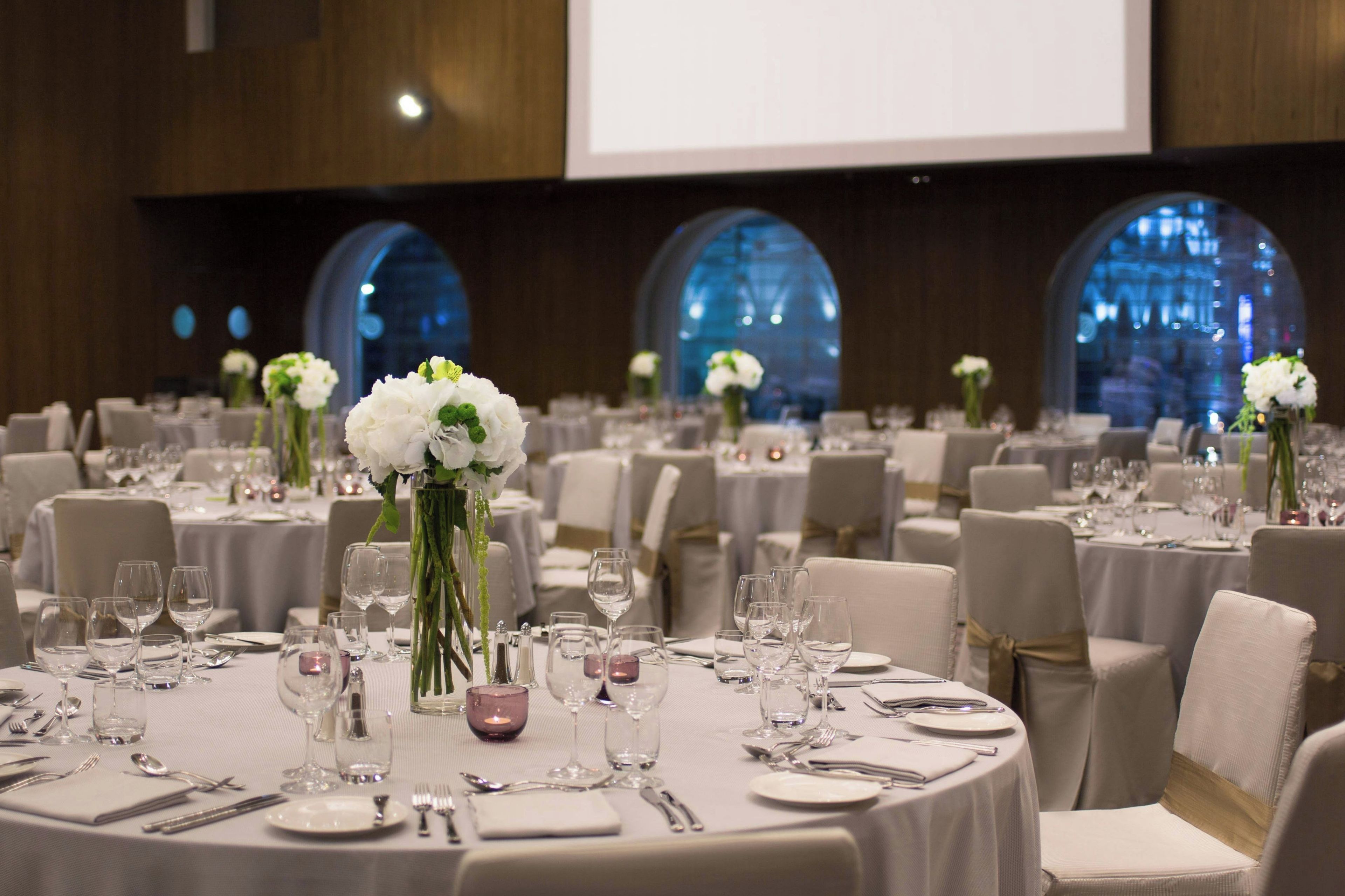 Une salle de banquet avec des tables rondes dressées pour le repas, chacune agrémentée d’un centre de table composé de fleurs blanches et de bougies.