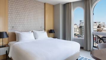 A hotel room with a large bed, two bedside tables with lamps, a view of the city through a balcony with curtains, and a statue on the balcony railing.