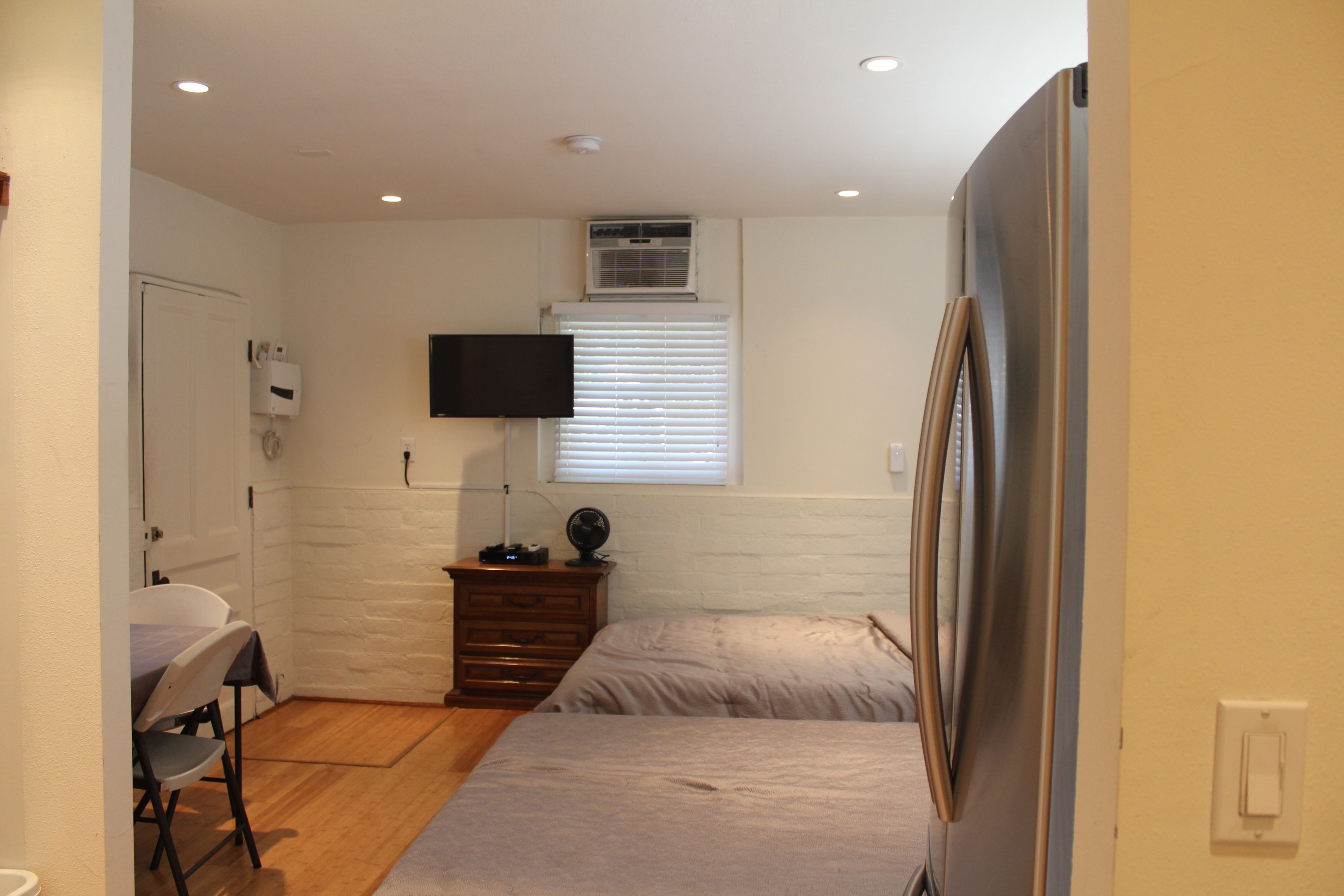 A compact bedroom with a single bed, a dresser, a desk with a chair, a television mounted on the wall, and a window with blinds.