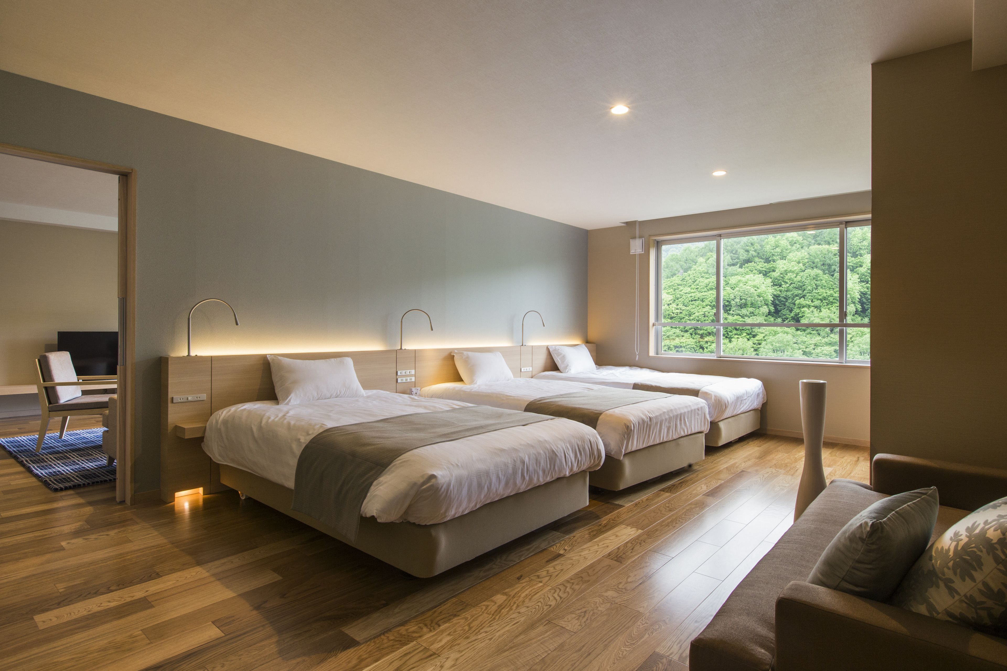 A modern hotel room with two beds, a large window, and a view of greenery.