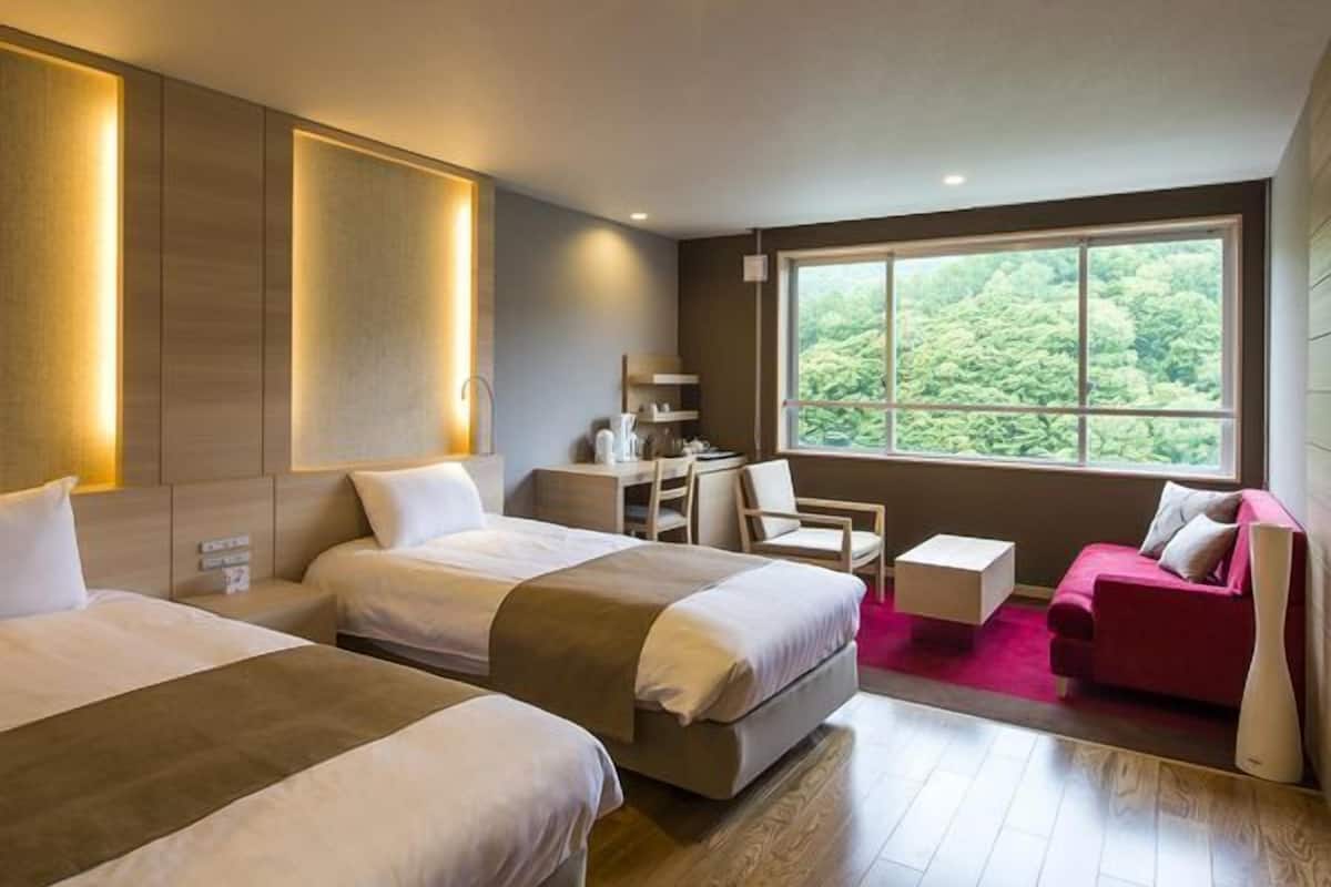 A hotel room with two beds, a desk, a chair, a small sofa, and a large window with a view of greenery.