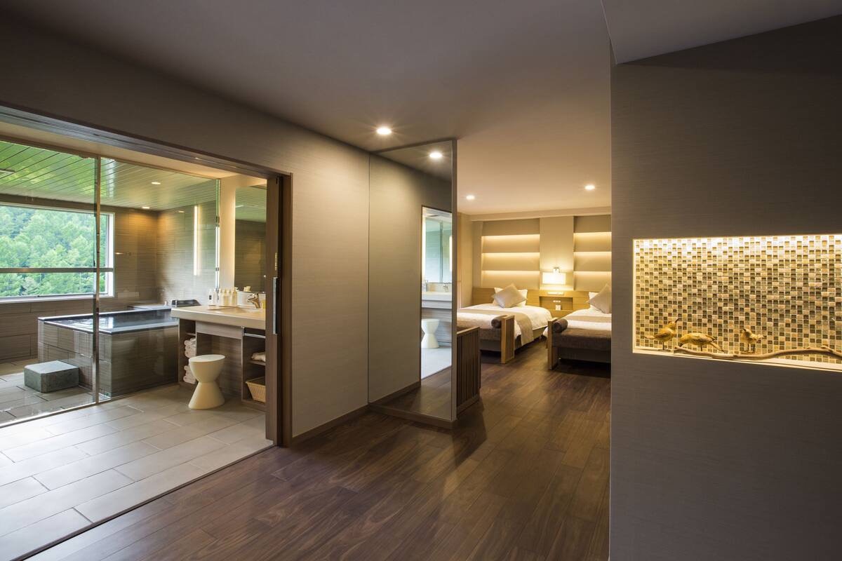 A modern hotel room with a glass shower enclosure, a wooden floor, a bed with a nightstand, and a decorative wall panel.