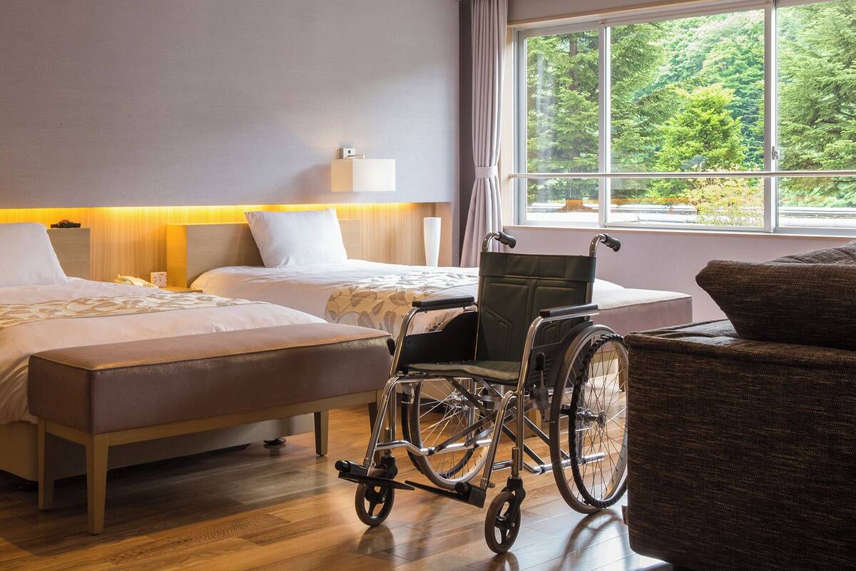 A hotel room with two beds, a wheelchair, and a view of greenery.