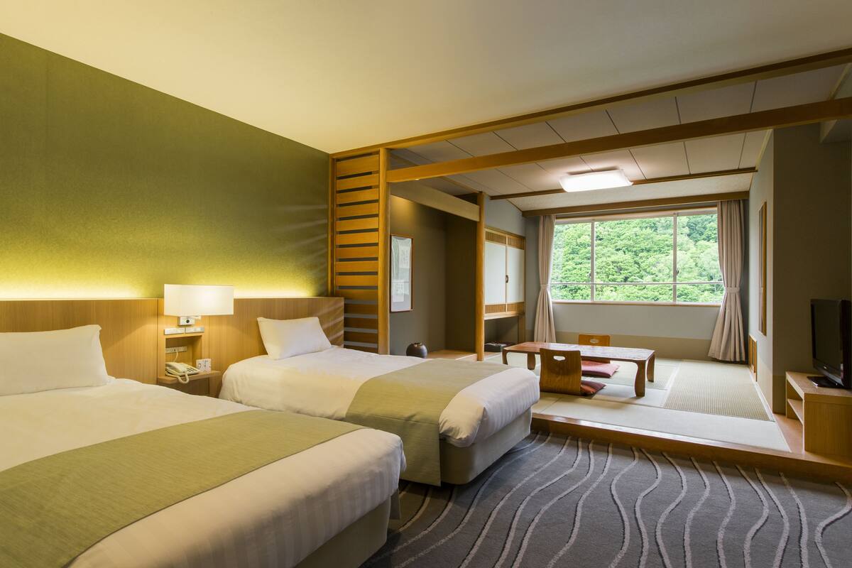 A hotel room with two beds, a desk, a chair, a TV, and a large window with a view of greenery.
