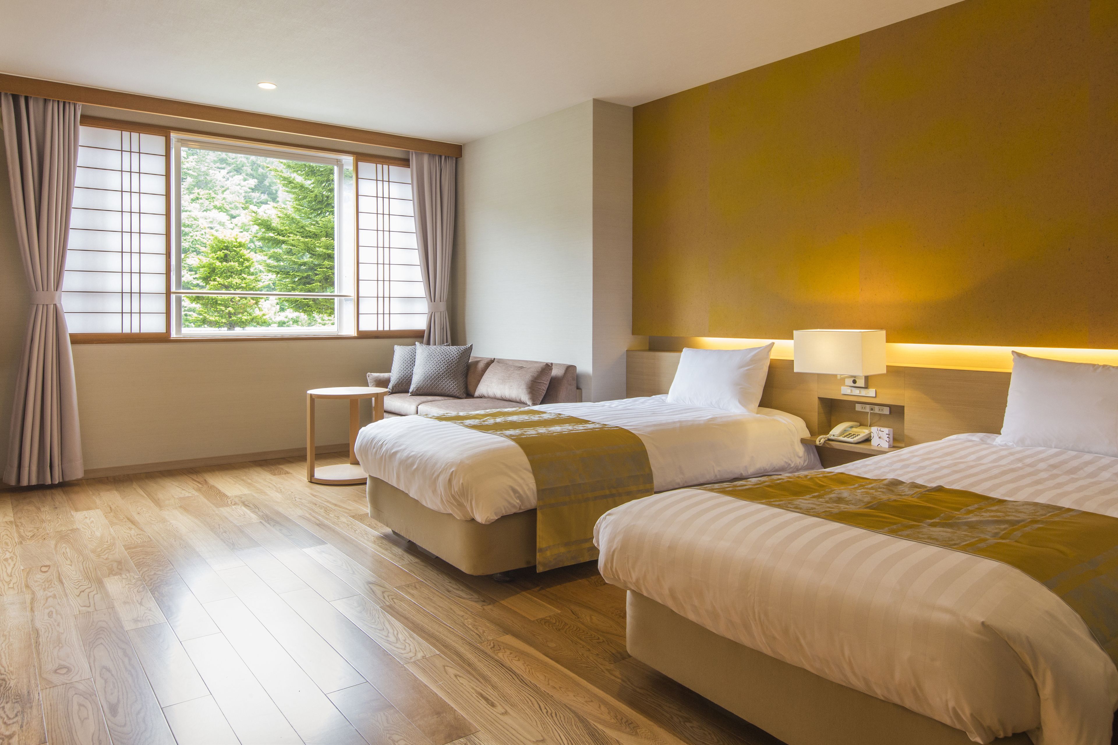 A hotel room with two beds, a window with curtains, a bedside table with a lamp, and a wooden floor.