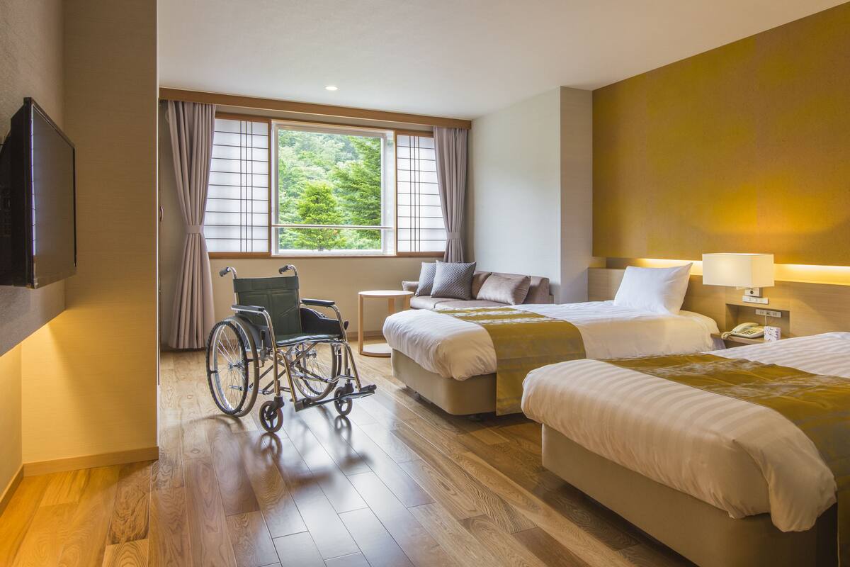 A hotel room with two beds, a wheelchair, a bedside table with a lamp, and a window with curtains.