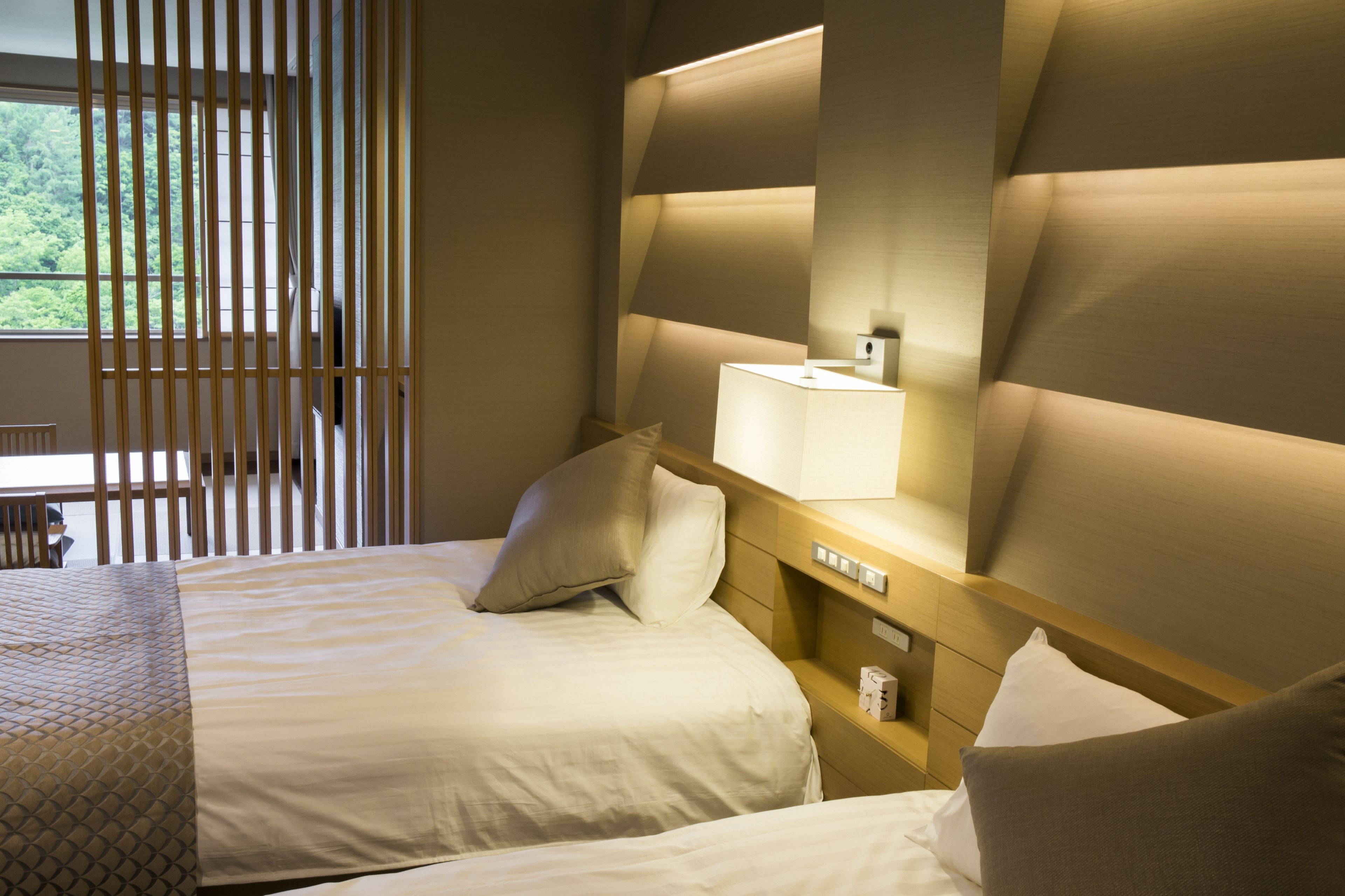 A hotel room with two beds, a nightstand, a lamp, and a view of greenery outside.